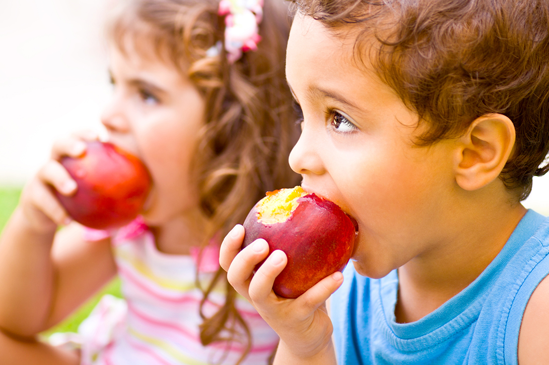 Photo of two happy children eating apples, brother and sister having picnic outdoors, cheerful kids biting red ripe peach, adorable infant holding fresh fruits in hands, healthy nutrition concept